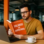 Homem jovem lendo o livro Falando em Empreender em um coworking moderno, com notebook e café sobre a mesa